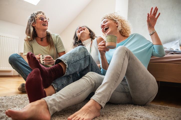 Three women in their 40s laughing together on the floor at home with mugs of coffee, illustrating the joy and health benefits of friendship in midlife