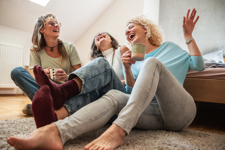 Three women in their 40s laughing together on the floor at home with mugs of coffee, illustrating the joy and health benefits of friendship in midlife